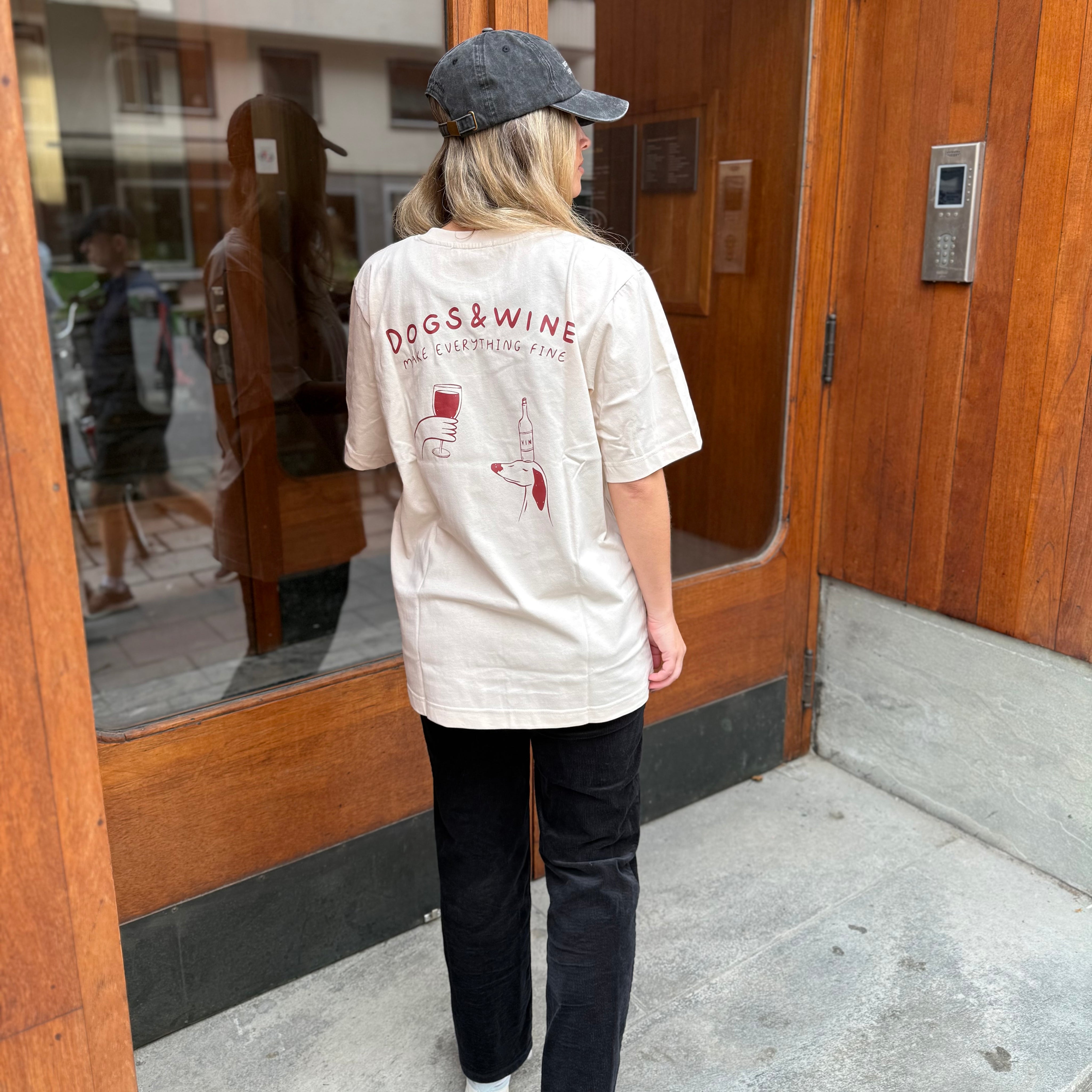 Person wearing a white t-shirt with 'DOGS & WINE' design, standing in front of a wooden door.