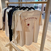 Off-white t-shirt with 'Cats & Wine' design on a wooden rack.
